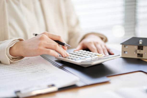 Personne calculant un budget de travaux avec une calculatrice, des documents administratifs et une maquette de maison